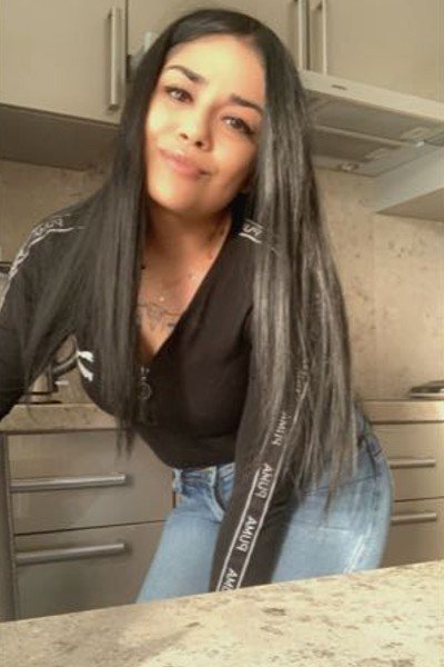 Rebecca neemt een selfie in haar keuken, gekleed in een blauwe spijkerbroek en een zwart shirt met lange mouwen. Ze lacht.